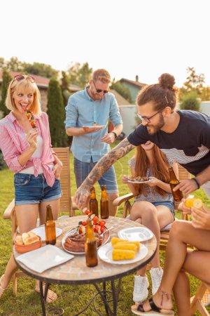 Téléchargez les photos : Groupe de jeunes amis joyeux réunis autour de la table, manger de la viande grillée, boire de la bière et s'amuser à une fête barbecue arrière-cour - en image libre de droit