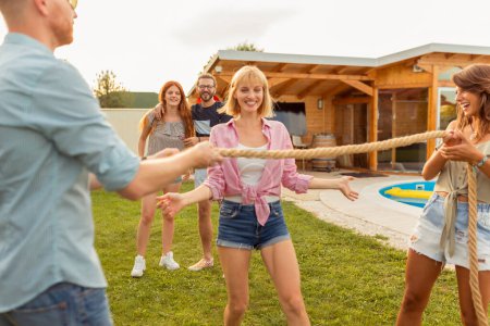 Foto de Grupo de jóvenes amigos alegres divirtiéndose en la fiesta al aire libre de verano junto a la piscina, haciendo el baile del limbo. Enfoque en la gente animando en el fondo - Imagen libre de derechos