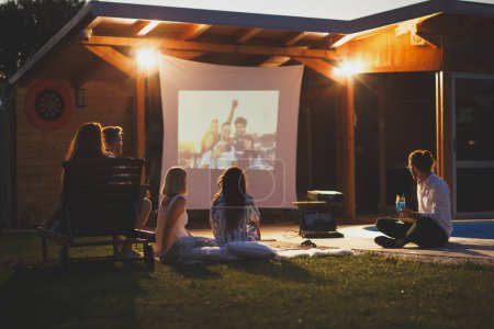 Téléchargez les photos : Groupe de jeunes amis joyeux qui s'amusent à manger du pop-corn, à boire des cocktails et à regarder un film dans un cinéma en plein air au bord de la piscine - en image libre de droit