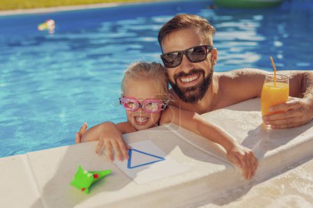 Téléchargez les photos : Belle petite fille et son père s'amusent à jouer à la piscine par une chaude journée ensoleillée d'été, père embrassant sa fille et buvant du jus d'orange - en image libre de droit