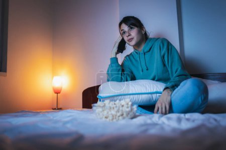 Téléchargez les photos : Belle jeune femme en pyjama assise au lit la nuit, mangeant du pop-corn et regardant un film à la télé - en image libre de droit