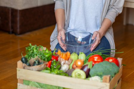 Photo for Detail of woman holding a bowl and sitting next to a wooden crate full of newly delivered fresh organic vegetables, picking out ingredients for cooking lunch - Royalty Free Image