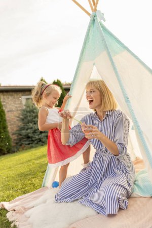 Foto de Hermosa madre e hija divirtiéndose acampando y jugando en el patio trasero, sentado en una tienda de campaña, haciendo y atrapando burbujas de jabón y relajándose al aire libre en un día soleado de verano - Imagen libre de derechos
