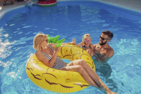 Téléchargez les photos : Les jeunes parents s'amusent à jouer avec leur fille dans la piscine, profiter d'une chaude journée ensoleillée en plein air et se détendre pendant les vacances d'été - en image libre de droit