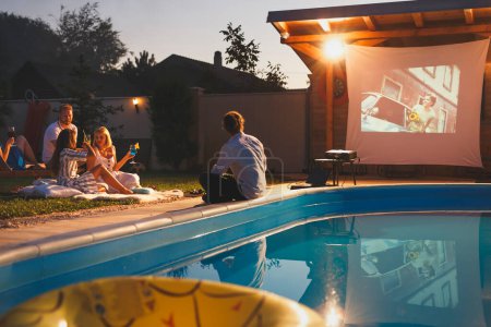 Téléchargez les photos : Groupe de jeunes amis joyeux qui s'amusent à manger du pop-corn, à boire des cocktails et à regarder un film dans un cinéma en plein air au bord de la piscine - en image libre de droit
