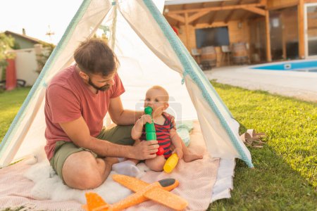 Foto de Joven padre divirtiéndose jugando con su bebé, acampando en el patio trasero junto a la piscina en un día soleado de verano - Imagen libre de derechos