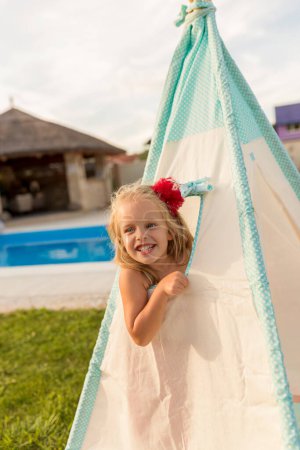 Foto de Hermosa niña alegre divirtiéndose mientras mira a través de una ventana de tienda mientras acampa en el patio trasero junto a la piscina - Imagen libre de derechos