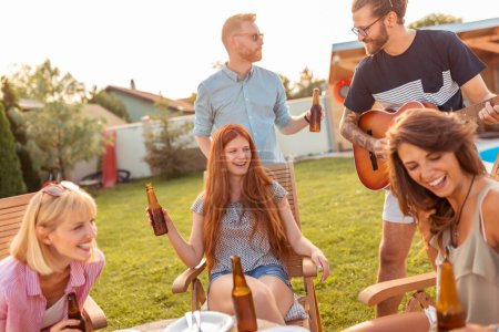 Foto de Grupo de amigos divirtiéndose tocando la guitarra y cantando en la fiesta de barbacoa del patio trasero, bebiendo cerveza y relajándose al aire libre en un día soleado de verano - Imagen libre de derechos