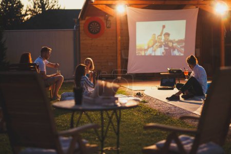 Photo for Group of cheerful young friends having fun eating popcorn, drinking cocktails and watching a movie in a home backyard poolside open air cinema - Royalty Free Image