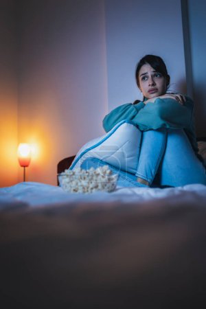 Téléchargez les photos : Belle jeune femme en pyjama assise au lit la nuit, triste en regardant un film dramatique à la télévision, étreignant un oreiller et pleurant - en image libre de droit