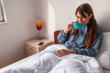 Téléchargez les photos : Belle jeune femme buvant son café du matin au lit après le réveil, profitant du temps libre à la maison et se relaxant - en image libre de droit