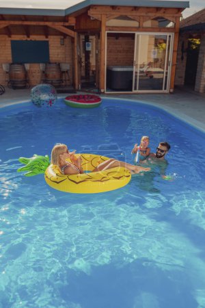 Téléchargez les photos : Les jeunes parents s'amusent à jouer avec leur fille dans la piscine, éclaboussant l'eau avec des pistolets à eau, profitant d'une chaude journée d'été ensoleillée à l'extérieur et se relaxant pendant les vacances d'été - en image libre de droit