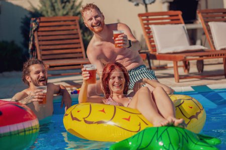 Grupo de amigos divirtiéndose levantando vasos de cerveza y haciendo un brindis en un caluroso día soleado de verano en la piscina, relajándose durante unas vacaciones de verano