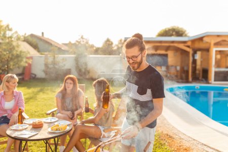 Téléchargez les photos : Groupe de jeunes amis faisant une fête barbecue dans la cour près de la piscine, homme grillant de la viande et buvant de la bière au premier plan pendant que les femmes sont assises et se relaxent en arrière-plan - en image libre de droit