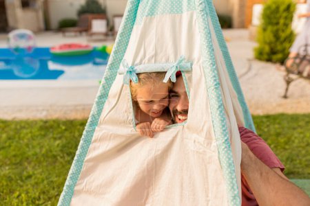 Photo for Father playing hide and seek with his little daughter while camping in the backyard by the swimming pool, having fun peeking out of the tent window while hiding - Royalty Free Image