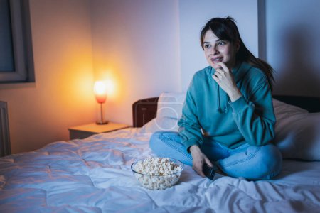Téléchargez les photos : Jeune femme en pyjama assis au lit, manger du pop-corn et rire en regardant un film drôle à la télévision, se détendre à la maison tard dans la nuit - en image libre de droit