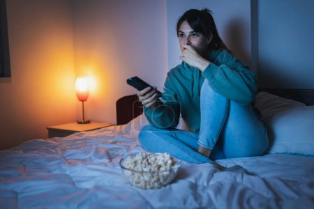 Téléchargez les photos : Jeune femme en pyjama assis au lit, manger du pop-corn et rire en regardant un film drôle à la télévision, changer de chaîne à l'aide de la télécommande - en image libre de droit