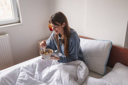 Schöne junge Frau im Schlafanzug sitzt morgens im Bett, isst Müsli mit frischen Beeren zum Frühstück und genießt die Freizeit zu Hause
