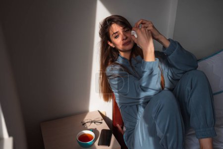Foto de Mujer joven deprimida usando pijamas, sentada en la cama por la mañana, limpiando lágrimas y nariz usando papel higiénico mientras llora - Imagen libre de derechos
