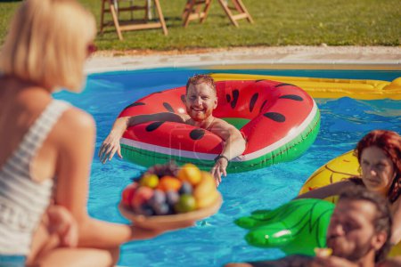 Téléchargez les photos : Groupe de jeunes amis s'amuser, se détendre et se rafraîchir dans la piscine par une chaude journée d'été ensoleillée, manger des fruits et flotter sur des matelas gonflables - en image libre de droit