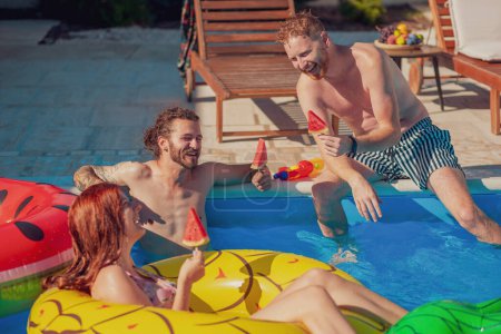 Foto de Grupo de jóvenes amigos divirtiéndose en la fiesta de verano en la piscina, pasando el soleado día de verano al aire libre y comiendo paletas de sandía junto a la piscina - Imagen libre de derechos
