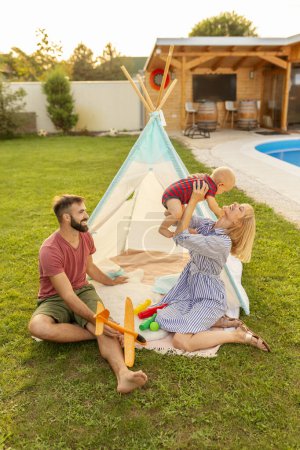 Foto de Padres jóvenes felices divirtiéndose acampando y jugando con su pequeño bebé junto a la piscina en el patio trasero de su casa, madre levantando a su hijo - Imagen libre de derechos
