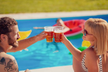 Foto de Hermosa pareja joven sentada junto a la piscina, tomando el sol y haciendo un brindis con vasos de cerveza mientras se relaja al aire libre en el soleado día de verano. - Imagen libre de derechos