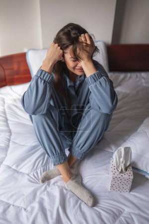 Photo for Desperate young woman wearing pajamas sitting on bed in the morning, holding head in hands, sobbing - Royalty Free Image