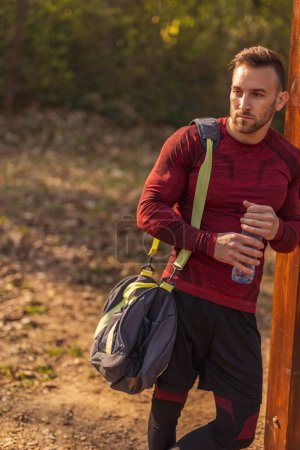 Photo for Athletic young man carrying gym bag, drinking water and relaxing after an intense outdoor workout in a street workout park - Royalty Free Image