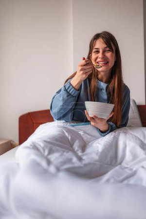 Schöne junge Frau im Schlafanzug sitzt morgens im Bett, isst Müsli mit frischen Beeren zum Frühstück und genießt die Freizeit zu Hause
