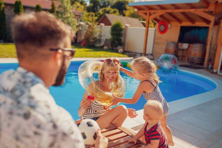 Photo for Parents playing with their little children by the swimming pool on a hot sunny summer day, little girl putting swim ring on her mother while baby boy is eating watermelon - Royalty Free Image