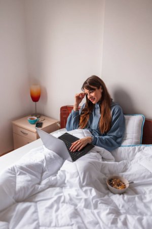 Photo for Young woman wearing pajamas sitting in bed in the morning, working from home using laptop computer and having breakfast in bed, eating cereal - Royalty Free Image