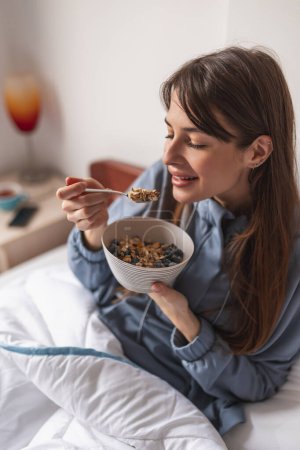 Hermosa joven con pijamas sentada en la cama por la mañana, tomando cereales con bayas frescas para el desayuno y disfrutando del tiempo libre en casa
