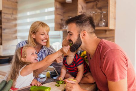 Photo for Beautiful happy family having fun cooking lunch together, sitting at kitchen counter, daughter feeding father with pieces of bell pepper cut for vegetable soup - Royalty Free Image
