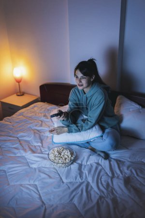 Téléchargez les photos : Vue en angle élevé d'une jeune femme s'amusant à regarder la télévision à la maison tard dans la nuit, changer de chaîne en utilisant la télécommande et manger du maïs soufflé - en image libre de droit