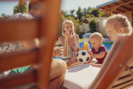 Foto de Hermosa familia joven pasar el día de verano soleado caliente junto a la piscina, los padres relajarse y beber cócteles mientras los niños están jugando en la cama de sol al lado de ellos - Imagen libre de derechos