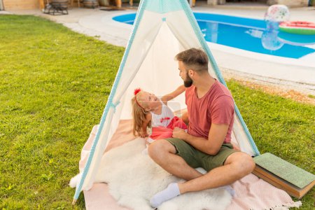 Téléchargez les photos : Père jouer avec sa petite fille tout en campant dans la cour près de la piscine, s'amuser assis dans la tente et passer des journées d'été ensoleillées en plein air - en image libre de droit