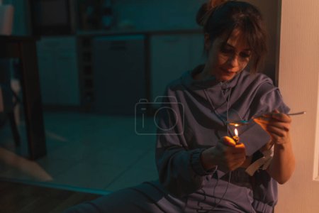 Photo for Female drug addict sitting on the floor in the dark, preparing next intravenous cocaine dose, using a lighter and a spoon; drug addiction and substance abuse concept - Royalty Free Image