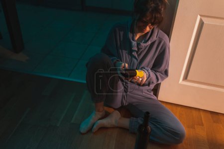 Photo for Female drug addict sitting on the kitchen floor in the dark drunk, taking a handful of pills with wine; substance abuse, depression and suicide concept - Royalty Free Image