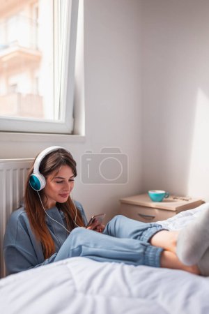 Téléchargez les photos : Femme assise sur le sol près du lit dans la chambre, se détendre à la maison le matin, porter un casque et apprendre la langue étrangère à l'aide de l'application de téléphone intelligent en ligne - en image libre de droit