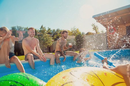 Téléchargez les photos : Groupe de jeunes amis joyeux s'amusant lors d'une fête d'été en plein air, assis près de la piscine, buvant de la bière et éclaboussant l'eau l'un sur l'autre - en image libre de droit