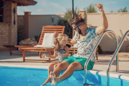 Téléchargez les photos : Beau père et fille joyeux assis au bord d'une piscine et s'amuser à l'extérieur par une chaude journée d'été ensoleillée - en image libre de droit