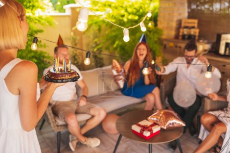 Téléchargez les photos : Groupe d'amis s'amuser à une fête d'anniversaire, chanter pendant que la fille d'anniversaire apporte gâteau - en image libre de droit