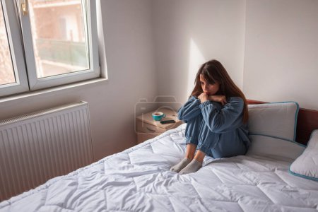 Depressive Frau im Schlafanzug sitzt morgens zusammengerollt und traurig auf dem Bett