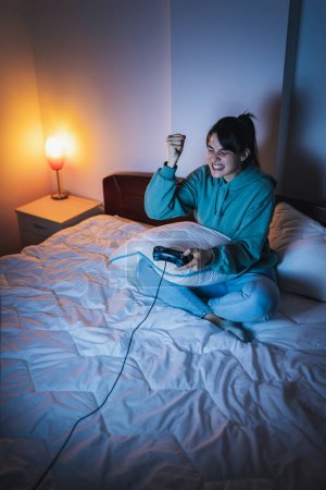 Téléchargez les photos : Belle jeune femme en pyjama s'amusant à la maison, jouant à des jeux vidéo au lit la nuit, excitée après avoir gagné le jeu - en image libre de droit
