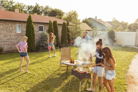 Téléchargez les photos : Groupe d'amis ayant une fête barbecue arrière-cour, griller de la viande, boire de la bière, jouer au frisbee et s'amuser par une journée d'été ensoleillée - en image libre de droit