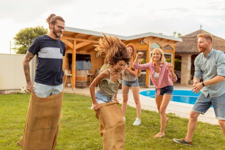 Foto de Grupo de alegres jóvenes amigos divirtiéndose mientras corren saltando en sacos en el patio trasero junto a la piscina - Imagen libre de derechos