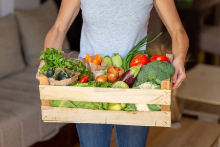 Téléchargez les photos : Femme portant une caisse en bois fraîchement livrée avec un large assortiment de légumes biologiques frais - en image libre de droit