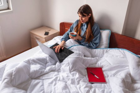 Téléchargez les photos : Belle jeune femme assise au lit le matin, travaillant de la maison à l'aide d'un ordinateur portable et buvant une tasse de thé chaud - en image libre de droit