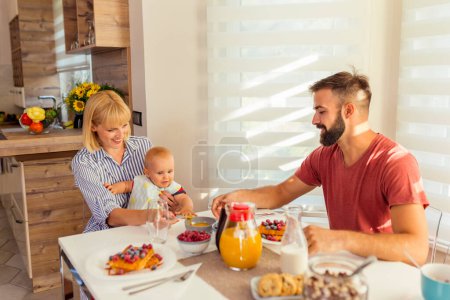 Téléchargez les photos : Les jeunes parents s'amusent à prendre le petit déjeuner et à nourrir leur petit garçon, appréciant passer le week-end matin ensemble à la maison - en image libre de droit
